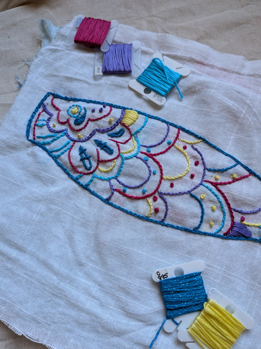 Broderie koinobori sur tissu lin écru — carpe japonaise aux écailles multicolores brodées au point de contour, avec fils DMC rose (600), violet (3746), turquoise (3846), bleu électrique (C995) et jaune (445) disposés autour du motif.